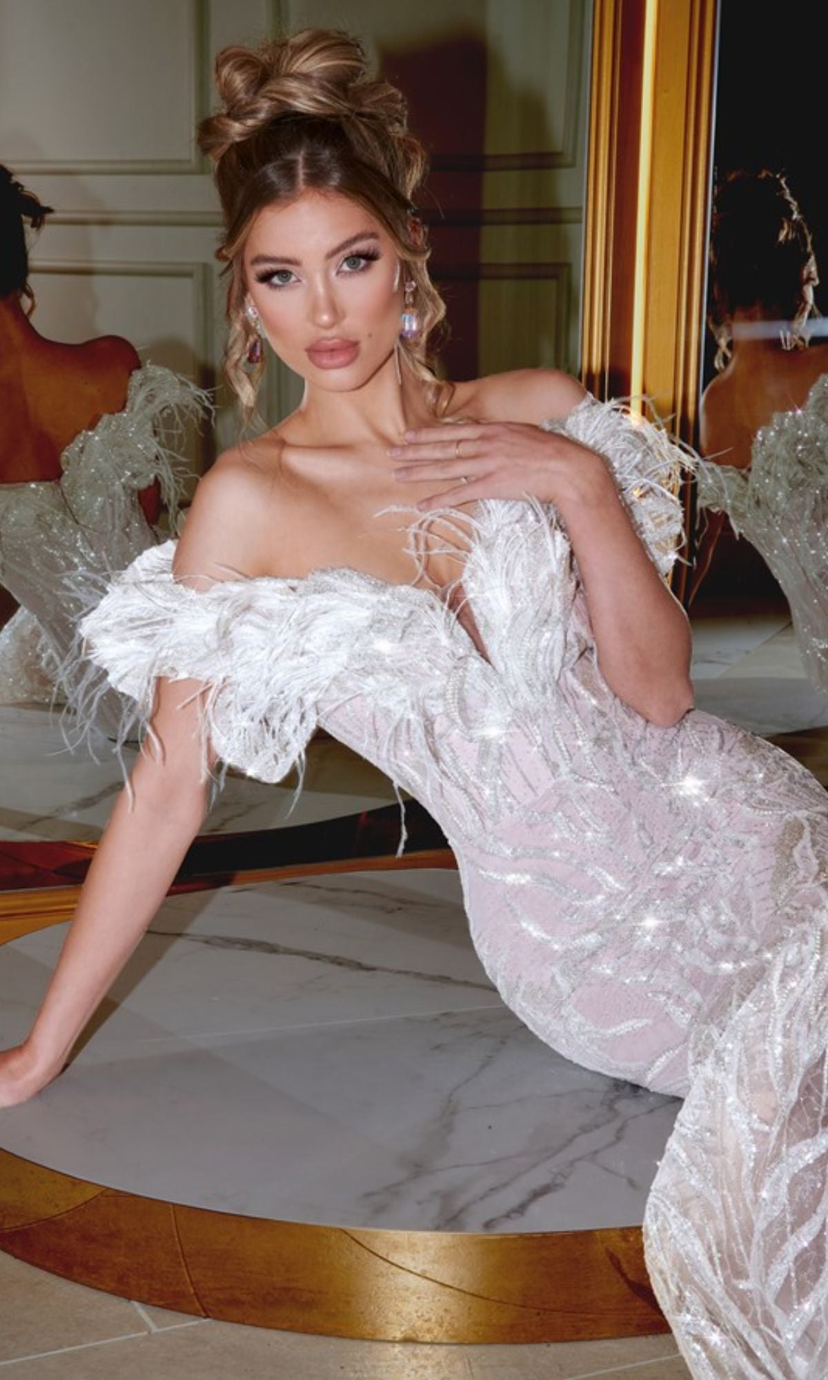 A woman poses on a marble floor in front of a mirror wearing the Long Prom Dress PS22227 by Portia and Scarlett, a white off-the-shoulder mermaid gown with feather trim.